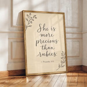 Könnte beinhalten: Ein rechteckiges gerahmtes Wandbild mit cremefarbenem Hintergrund. Der Text "She is more precious than rubies. - Proverbs 3:15" ist in schwarzer Schreibschrift gedruckt. Zarte schwarze Strichzeichnungen von Blumenranken zieren die Ecken.