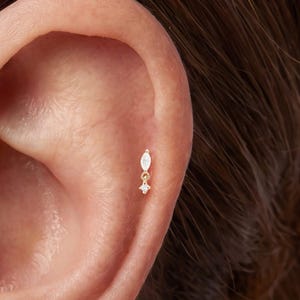 May include: Two gold-toned stud earrings featuring a marquise-cut clear stone positioned above a smaller, four-pointed star-shaped clear stone. One earring is placed in the tragus, and the other is positioned in the upper cartilage of the ear.
