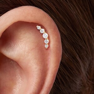 May include: A gold cartilage earring featuring a curved row of five round, clear, prong-set stones that graduate in size. The jewelry is positioned along the upper edge of the ear, following the natural curve of the cartilage with a metallic finish.