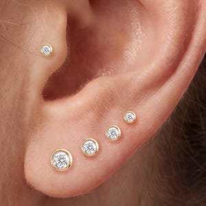 Peut inclure: Une collection de boucles d'oreilles à clous en or avec des pierres rondes transparentes, comprenant trois clous sur le cartilage supérieur, un dans le rook, un dans le tragus et quatre clous gradués sur le lobe en disposition verticale.