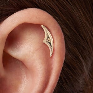 Könnte beinhalten: Ein goldfarbenes Knorpel-Piercing für das Ohr in einer geschwungenen, spitz zulaufenden, flügelartigen Form mit einem kleinen, eingravierten Triquetra-Knotensymbol in der Mitte des Designs, platziert am oberen Rand des Ohrknorpels.