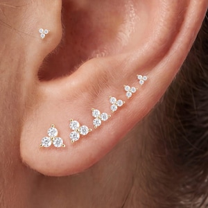 May include: A collection of gold stud earrings, each featuring three small, clear, round-cut stones arranged in a triangular cluster. These earrings are placed in multiple piercings along the earlobe and the upper cartilage of the ear.