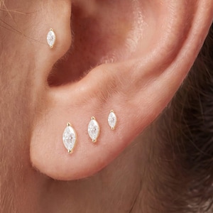 May include: A set of four gold-toned stud earrings, each featuring a marquise-cut clear gemstone. Three studs are arranged along the earlobe in descending size, while the fourth stud is positioned in the upper cartilage area of the ear.