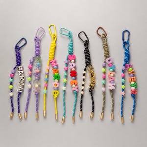 May include: Eight colorful paracord keychains with metal carabiners and gold-tone end caps. Each keychain features various plastic letter beads spelling names like EARIN, MARCO, SANEY, NORA, DYLAN, and AVERY, accented with small star and round beads.