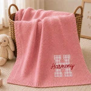 May include: A soft pink knitted blanket featuring a large white and pink gingham letter H with the name Harmony embroidered in red script across the centre. The blanket has a ribbed border and is draped over a woven basket next to a plush toy.