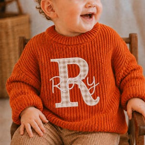 May include: A vibrant orange long-sleeve knit sweater featuring a large, beige and white gingham-patterned letter R appliqued on the front. The name "Rory" is embroidered in white cursive script across the center of the letter R.