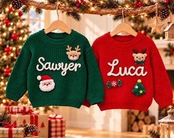 Op de afbeelding: Twee gebreide truien met lange mouwen aan houten kledinghangers. De groene trui heeft de naam Sawyer in witte letters, een pluche kerstmangezicht en een rendier. De rode trui heeft de naam Luca, een rendier, een snoepje en een kerstboom.
