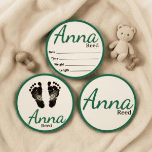 May include: Three circular wooden plaques with dark green borders. One plaque features the name Anna Reed with lines for date, time, weight, and length. Another displays black baby footprints and the name Anna Reed. The third shows the name Anna Reed.