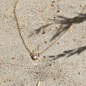 May include: A delicate gold necklace with a small, round, clear gemstone pendant. The necklace is lying on a textured, light gray surface, with shadows from foliage in the background.