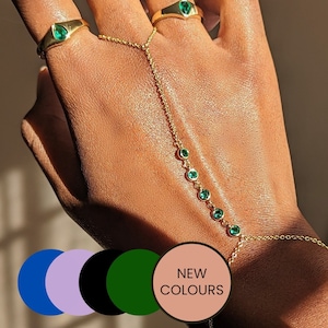 Peut inclure: Un bracelet en chaîne dorée avec une pierre verte émeraude en forme de larme sur chaque maillon. Le bracelet est porté sur une main avec deux anneaux en or, chacun avec une seule pierre verte émeraude.