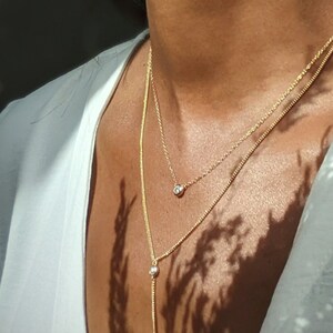 May include: A gold-colored necklace with two strands, one short and one long, each adorned with small, clear gemstones. The necklace is layered and worn against a white top, with shadows from foliage adding contrast.
