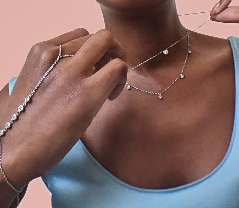 May include: A silver necklace and hand chain set with clear, round gemstones. The necklace has multiple dangling stones. The hand chain connects to a ring and features a series of gemstones. The jewelry is displayed against a light blue top and a pink background.