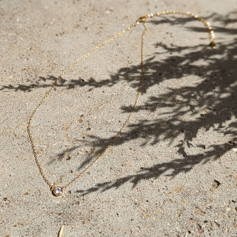 May include: A delicate gold necklace with a small, round, clear gemstone pendant. The necklace is lying on a textured, light gray surface, with shadows from foliage in the background.