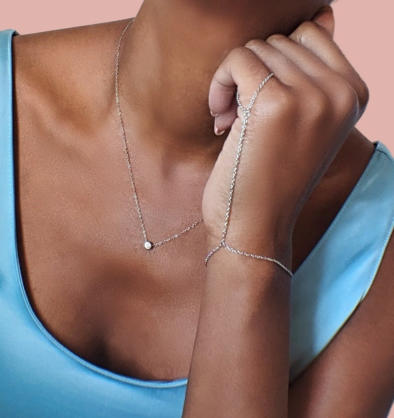 May include: A delicate silver necklace with a small, round pendant and a matching hand chain bracelet. The jewelry is displayed against a light blue background, creating a minimalist and elegant aesthetic. The jewelry is designed to be worn together.