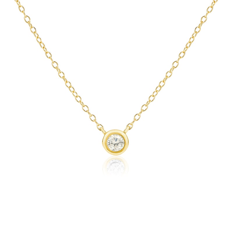 May include: A delicate gold necklace featuring a single, round, clear gemstone pendant. The pendant is set in a bezel setting and hangs from a fine gold chain. The necklace is displayed against a white background.