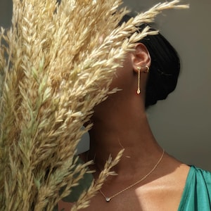 May include: A gold necklace with a small, round, clear gemstone pendant. A matching pair of gold earrings with a dangling teardrop-shaped charm are visible. The jewelry is worn against a dark background, with a green top and dried wheat in the foreground.