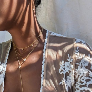 May include: A layered gold necklace with three strands, each featuring small, sparkling gemstones. The necklace is paired with matching gold earrings. The model is wearing a sheer, cream-colored top with white floral embroidery.