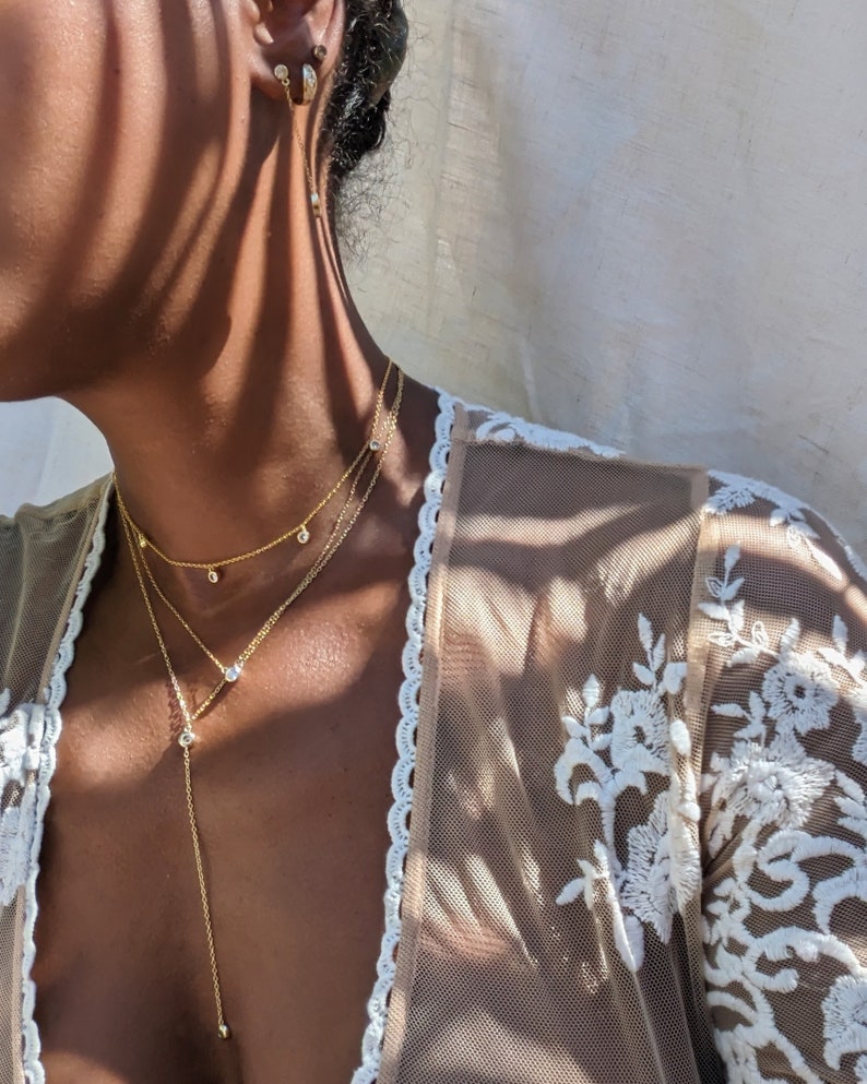 May include: A layered gold necklace with three strands, each featuring small, sparkling gemstones. The necklace is paired with matching gold earrings. The model is wearing a sheer, cream-colored top with white floral embroidery.