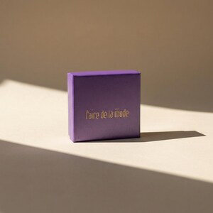May include: A small, square, purple box with the phrase "faire de la mode" printed in gold lettering. The box is set against a neutral background with a shadow cast across the surface.