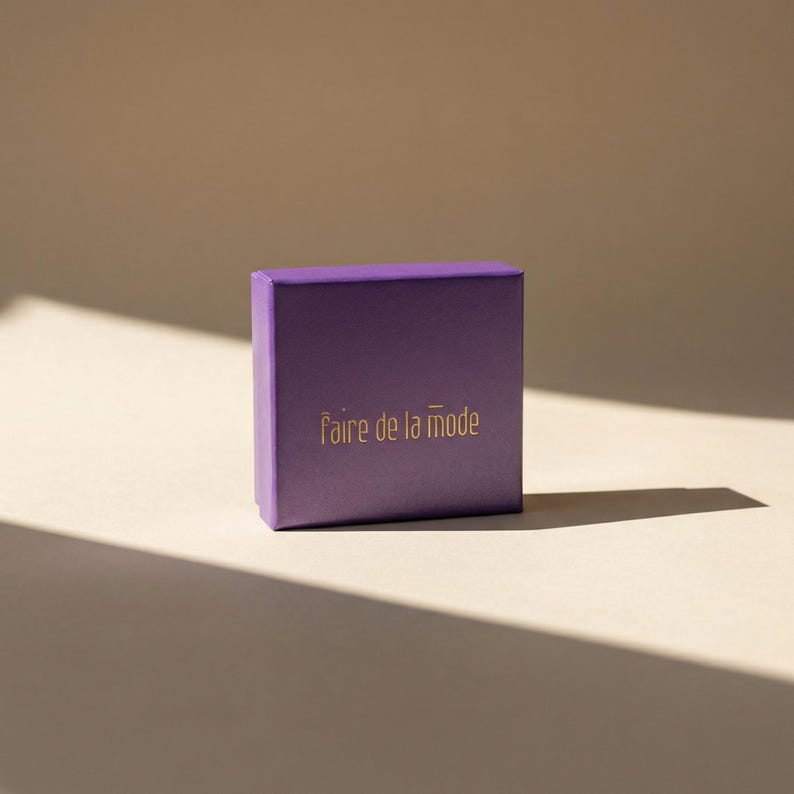 May include: A small, square, purple box with the phrase "faire de la mode" printed in gold lettering. The box is set against a neutral background with a shadow cast across the surface.