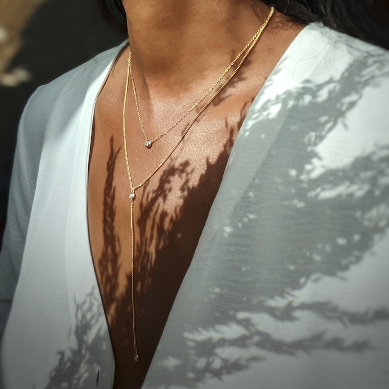 May include: A gold-colored necklace with two strands, one short and one long, each adorned with small, clear gemstones. The necklace is layered and worn against a white top, with shadows from foliage adding contrast.