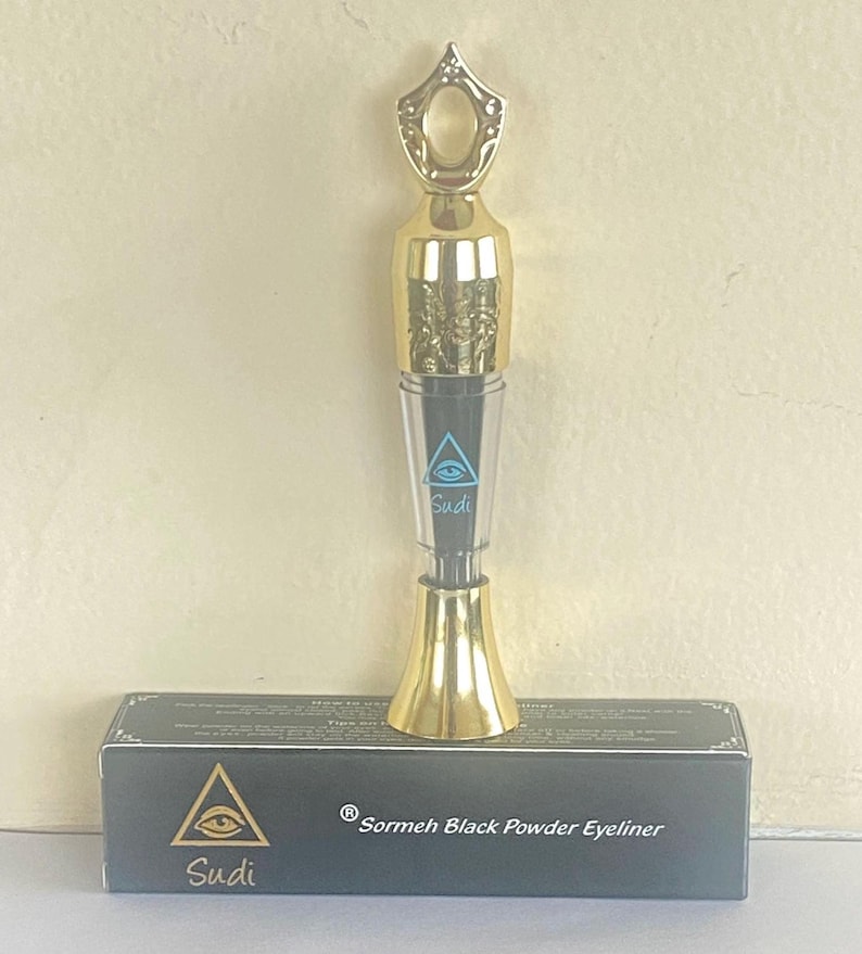 May include: A gold-colored eyeliner applicator with a decorative top and a clear tube. The applicator is labeled "Sudi" and sits atop a black box with the text "Sudi" and "Sormeh Black Powder Eyeliner".