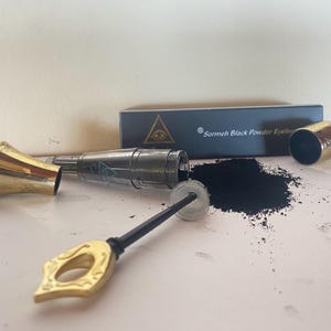以下が含まれることがあります： 金と黒の容器に入った黒いパウダーアイライナー。小さな金のロートと黒いパウダーアプリケーターが付いています。容器には「Sormeh Black Powder Eyeliner」の文字が書かれています。