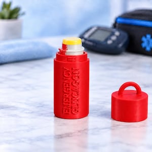 May include: A bright red cylindrical emergency glucagon case with a matching red cap featuring a small loop on top. The case has the words EMERGENCY GLUCAGON embossed vertically on the side, and it is shown open with a yellow and white inner component.