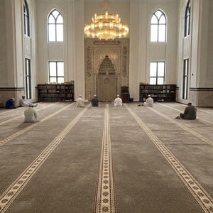 May include: A spacious mosque interior featuring a large, beige prayer carpet with linear patterns, a central ornate mihrab, and a multi-tiered gold chandelier. Several individuals are seated on the floor in prayer, with tall arched windows and wooden bookshelves lining the white walls.