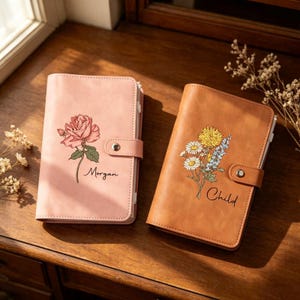 May include: Two rectangular notebooks with snap closures. The left notebook is light pink with a rose illustration and the name Morgan. The right notebook is tan with a wildflower bouquet illustration and the name Child. Both have smooth covers.
