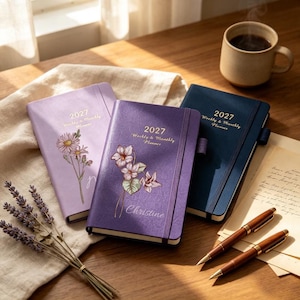 May include: Three 2027 weekly and monthly planners in light purple, dark purple, and navy blue. Each planner features a floral illustration on the cover, an elastic closure band, and gold text reading 2027 Weekly & Monthly Planner.