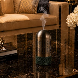 Peut inclure: Un brûle-encens cylindrique doté d'un dôme supérieur en métal bronze avec des découpes verticales et d'une base en marbre vert massif. Un mince filet de fumée blanche s'élève du centre du dôme, indiquant l'utilisation active de l'objet.