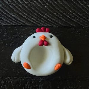 May include: A round, white clay chicken figurine with a smooth finish. The chicken features a small orange beak, two dark red wattles, a red comb, two black eyes, and two small orange feet. The center of the chicken body has a circular indentation.