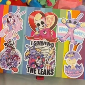 Op de afbeelding: Een rechthoekige doos met verticale regenboogstrepen bedekt met diverse karakterstickers, waaronder een met de tekst I SURVIVED THE LEAKS, geplaatst voor twee knuffels, een paars-rode nar en een paarse konijnachtige figuur.