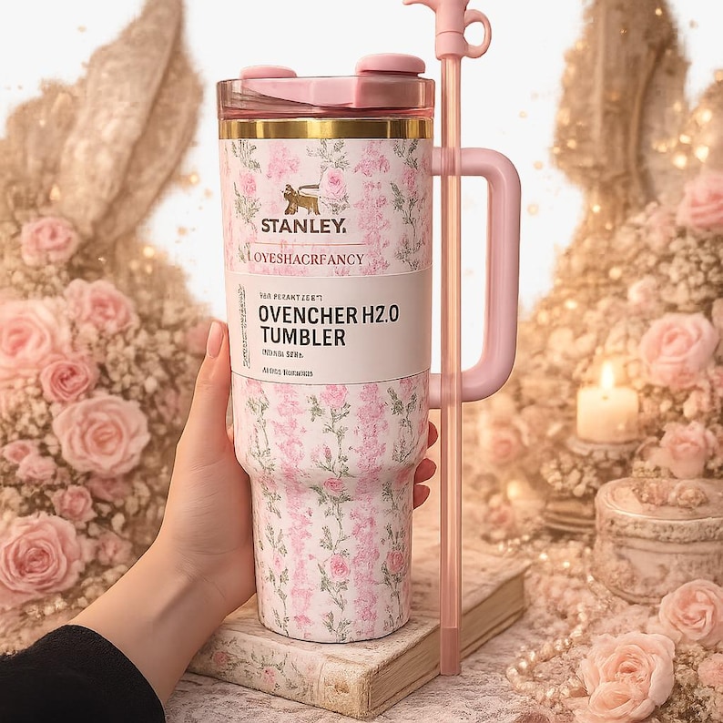 May include: A Stanley LoveShackFancy Quencher H2.0 tumbler featuring a pink floral pattern on a white background, a pink handle, a matching pink lid, and a long pink straw. The tumbler has gold-tone rim accents and clear branding text.