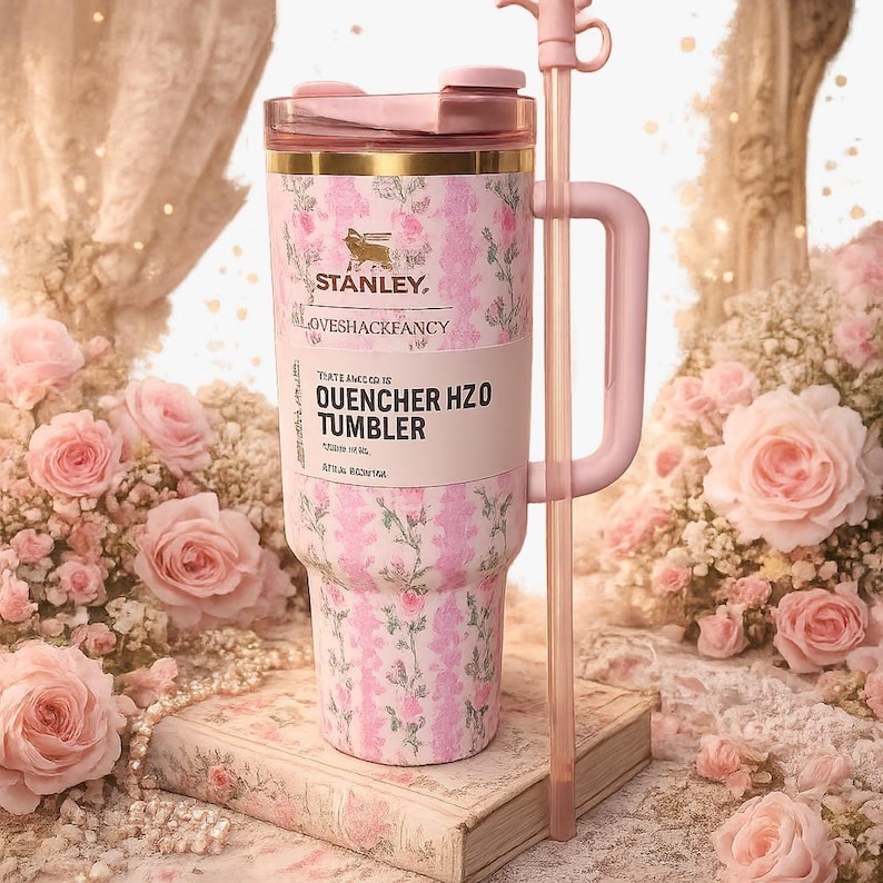 May include: A pink and white floral patterned Stanley LoveShackFancy Quencher H2O tumbler with a matching pink handle, a clear lid, and a long pink straw. The tumbler features a gold rim and the text Stanley LoveShackFancy Quencher H2O Tumbler.