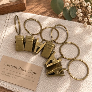 Puede incluir: Un conjunto de clips con anillas para cortinas de metal en tono latón antiguo. Cada clip cuenta con una anilla circular unida a una pinza rectangular con resorte. Se incluye una pequeña tarjeta rectangular con el texto: Curtain Ring Clips, FOR CURTAINS / FABRICS / HOME DECOR, FOR EVERYDAY USE.