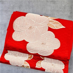 May include: A folded red textile featuring a woven pattern of large, pale beige camellia flowers and leaves. The fabric has a subtle, repeating diamond-shaped texture across the vibrant red background, creating a layered and detailed appearance.