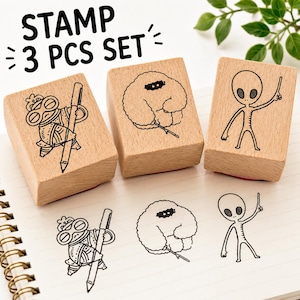 May include: A set of three wooden stamps labelled STAMP 3 PCS SET. Each stamp features a unique black ink design: a character wearing goggles holding a large pencil, a fluffy creature holding a stick, and an alien figure pointing upwards.