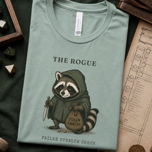 Może przedstawiać: Szałwiowo-zielony t-shirt z grafiką szopa pracza w ciemnozielonym płaszczu z kapturem. Szop trzyma sztylet i worek z napisem "STOLEN SNACKS". Powyżej znajduje się napis "THE ROGUE", a poniżej "FAILED STEALTH CHECK".
