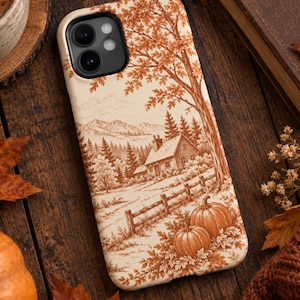 May include: A cream-colored phone case featuring a detailed monochromatic burnt orange illustration of a rustic cabin nestled in a forest with mountains in the background, a wooden fence, and two pumpkins resting in the foreground among fallen leaves.
