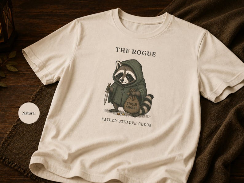 Może przedstawiać: T-shirt w naturalnym kolorze z grafiką szopa pracza w zielonym kapturze, trzymającego mały sztylet i worek z napisem STOLEN SNACKS. Nad szopem znajduje się napis THE ROGUE, a pod nim FAILED STEALTH CHECK.