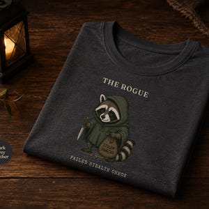 Może przedstawiać: Ciemnoszary melanżowy t-shirt z grafiką szopa pracza w zielonej pelerynie z kapturem, trzymającego mały n&oacute;ż i worek z napisem STOLEN SNACKS. Powyżej znajduje się napis THE ROGUE, a poniżej napis FAILED STEALTH CHECK.