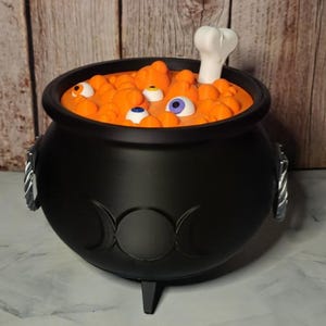 May include: A black cauldron with a triple moon symbol on the front and silver-colored side handles. The interior is filled with orange bubbles, several multi-colored eyeballs, and a single white bone protruding from the top surface.