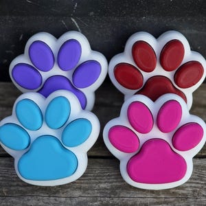 May include: Four paw-shaped objects with white borders and colored centers. One has purple pads, one has red pads, one has light blue pads, and one has hot pink pads. Each object features a central large pad and four smaller oval pads arranged in a paw print.