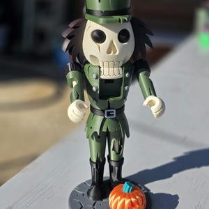 May include: A nutcracker figurine featuring a skeletal head with dark hair, wearing a green military-style uniform with a matching hat. The figure stands on a circular gray base next to a small orange pumpkin with a blue stem.