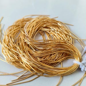 Peut inclure: Un faisceau de fils métalliques fins et flexibles de couleur dorée, enroulés en forme circulaire. Le fil métallique présente une surface lisse et réfléchissante, maintenu à une extrémité par un petit morceau de ficelle blanche, avec des extrémités libres.