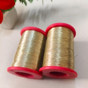 Pode incluir: Dois carretéis cilíndricos de fio metálico dourado, cada um com tampas de plástico vermelho brilhante. O fio é enrolado firmemente em torno do centro dos carretéis, criando uma textura de superfície cintilante e reflexiva nas fibras douradas.