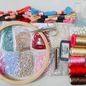 May include: A craft supply kit featuring a wooden embroidery hoop, metal scissors, and various embroidery threads in red, pink, blue, and gold. Includes multiple packets of sequins, beads, and pearls, along with needles and spools of metallic thread.