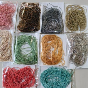 May include: Ten individual clear plastic bags containing coiled metallic and solid colored craft cords. The colors include pink, dark brown, charcoal gray, gold, beige, light green, orange, silver, red, and teal, arranged in a grid-like formation.